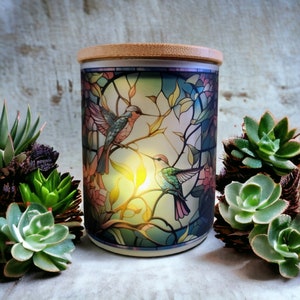 May include: A ceramic candle jar with a stained glass design featuring two hummingbirds in flight. The jar has a wooden lid and is surrounded by succulents.