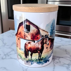 May include: A white ceramic canister with a wooden lid, featuring a watercolor painting of a brown horse standing in front of a red barn. The design includes blue and green accents, creating a rustic aesthetic.