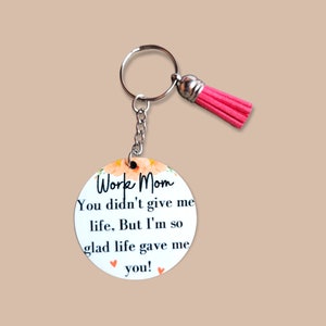 May include: A round keychain with a floral design and the text "Work Mom You didn't give me life, But I'm so glad life gave me you!" with two pink hearts. The keychain has a silver ring and a pink tassel.
