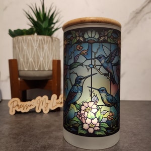 May include: A white ceramic candle holder with a stained glass design featuring three blue hummingbirds and pink flowers. The candle holder has a bamboo lid.