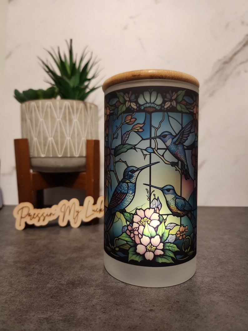 May include: A white ceramic candle holder with a stained glass design featuring three blue hummingbirds and pink flowers. The candle holder has a bamboo lid.