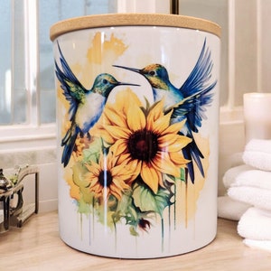 Puede incluir: Un tarro de cerámica blanco con tapa de bambú que presenta un diseño de acuarela de dos colibríes flotando alrededor de un girasol con pétalos amarillos y un centro marrón. El fondo es de color amarillo claro con salpicaduras de acuarela.