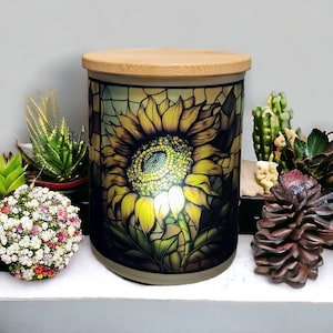 May include: A ceramic jar with a wooden lid features a stained glass design of a sunflower with yellow petals and a dark brown center. The jar is illuminated from within, creating a warm glow.