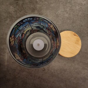 May include: A glass candle holder with a stained glass design featuring birds and flowers. The holder has a clear glass interior and a wooden lid.