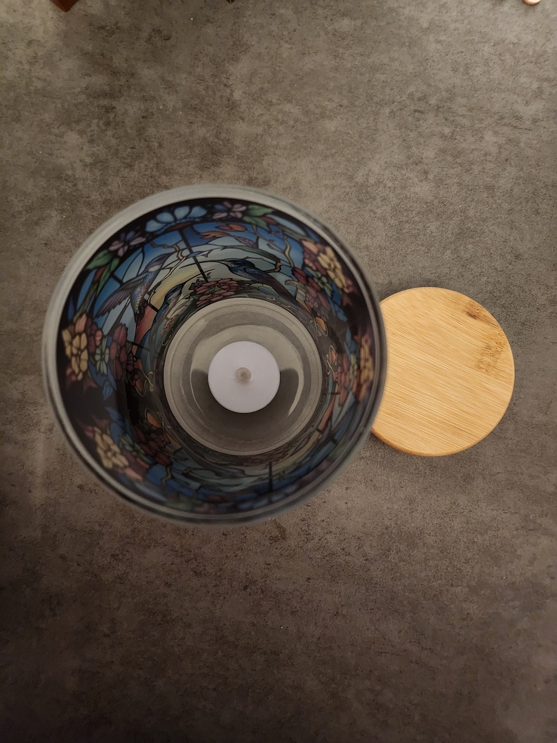 May include: A glass candle holder with a stained glass design featuring birds and flowers. The holder has a clear glass interior and a wooden lid.