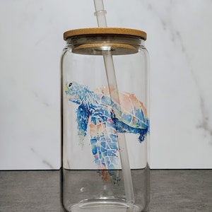 May include: A clear glass tumbler with a bamboo lid and a clear straw. The tumbler features a watercolor design of a sea turtle in shades of blue, orange, and pink. The tumbler is designed for cold beverages.