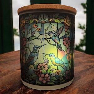 May include: A ceramic jar with a wooden lid features a stained glass design of three hummingbirds in flight. The hummingbirds are blue, green, and yellow with black outlines. The jar is surrounded by pink and white flowers.