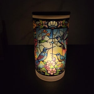 May include: A white ceramic candle holder with a stained glass design featuring three blue hummingbirds and pink flowers. The candle holder is lit and glowing.