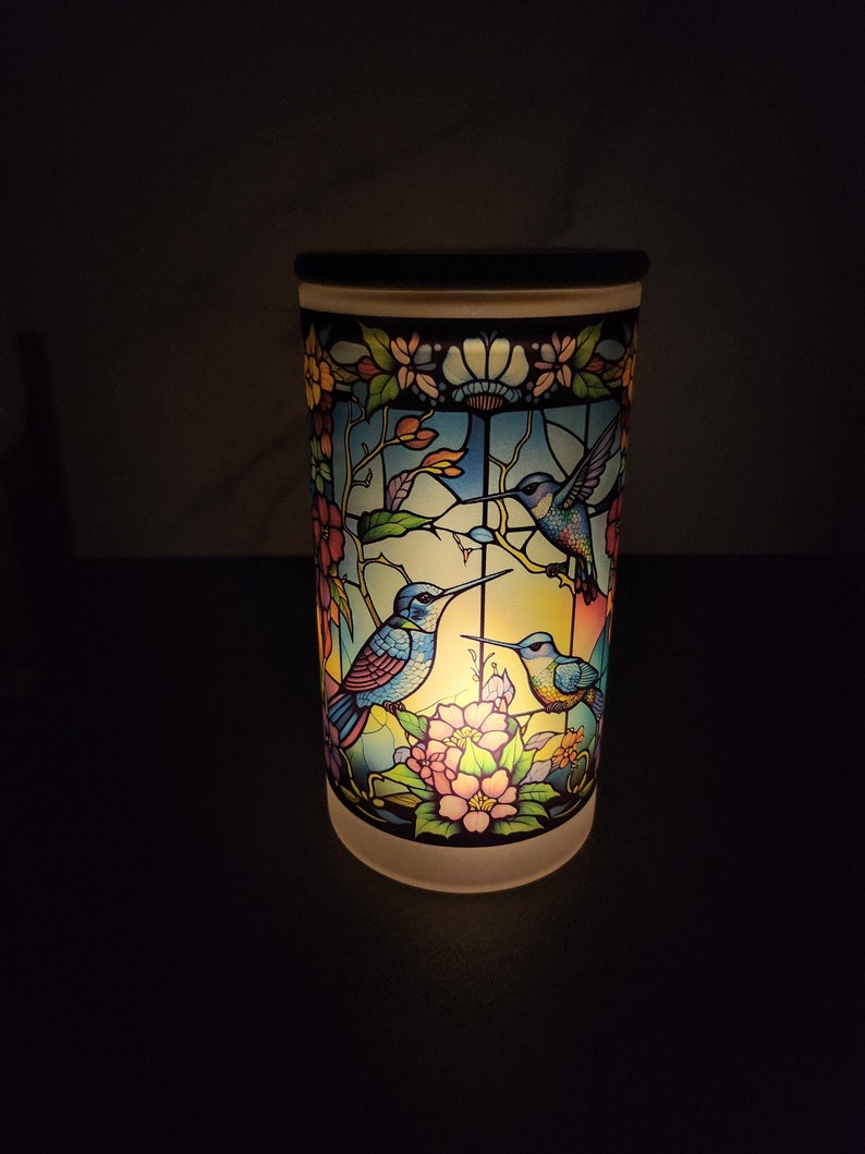 May include: A white ceramic candle holder with a stained glass design featuring three blue hummingbirds and pink flowers. The candle holder is lit and glowing.