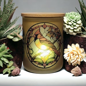 May include: A glass candle jar with a wooden lid features a stained glass design of a cardinal bird perched on a branch. The design is in shades of red, yellow, green, and brown.
