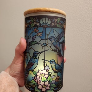May include: A cylindrical glass jar with a wooden lid, featuring a stained glass design of three hummingbirds and pink flowers. The jar is illuminated from within, creating a warm glow.