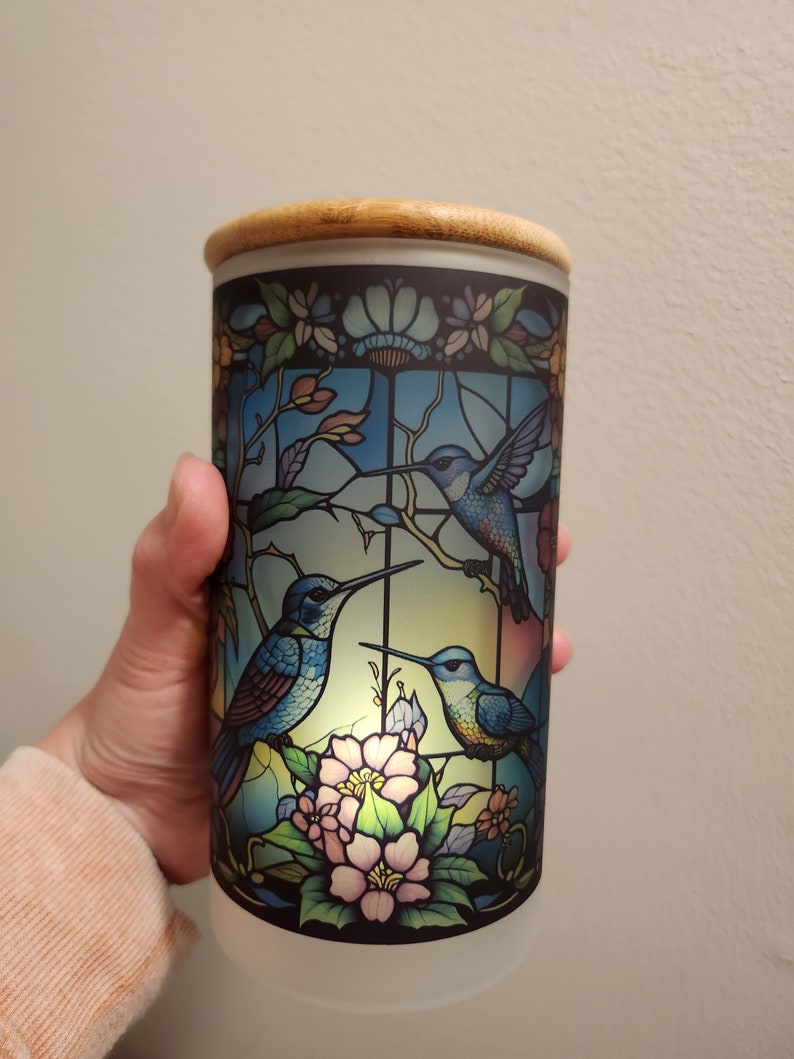 May include: A cylindrical glass jar with a wooden lid, featuring a stained glass design of three hummingbirds and pink flowers. The jar is illuminated from within, creating a warm glow.