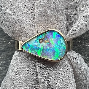 May include: A gold ring featuring a pear-shaped opal with vibrant blue, green, and white hues. The opal is set in a gold bezel, creating a striking contrast. The ring is displayed on a gray, textured fabric.