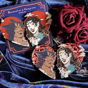 Op de afbeelding: Twee emaillepins die een weerwolf en een vampier voorstellen, elk met een halve hartvorm. De weerwolf is bruin en zwart met een tandenrij, en de vampir is een vrouw met lang donker haar en een rode jurk. De pins zijn op een blauwe fluwelen achtergrond met een rode roos.