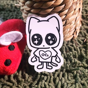 May include: A white sticker featuring a cartoon cat with large, sparkling eyes and a heart-shaped shirt with the letters "OCs". The sticker is outlined in black and has a simple, cute design. A red plush toy and woven basket are in the background.
