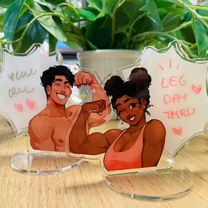 以下が含まれることがあります： 吹き出し付きのアクリル製キャラクタースタンド2つ。左側のスタンドは茶色の髪をした男性で、茶色と白の吹き出しに「You bet」と書かれています。右側のスタンドは茶色の髪をした女性で、ピンクと白の吹き出しに「Leg day tmrw」と書かれています。