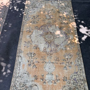 Puede incluir: Una alfombra vintage con fondo beige y un patrón floral descolorido en tonos marrones, morados y blancos. La alfombra tiene un aspecto desgastado y un borde con flecos.