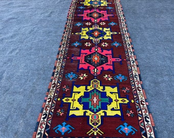 Op de afbeelding: Een lang, smal tapijt met een traditioneel geometrisch patroon in tinten rood, blauw, geel en wit. Het tapijt heeft een donkerbruine achtergrond en een rand met een herhalend patroon.