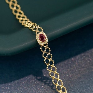 May include: A gold chain bracelet featuring a red gemstone set in a halo of smaller white gemstones.