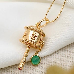 May include: Gold necklace with a spinning prayer wheel pendant. The pendant has intricate details and a small green gemstone hanging from it.