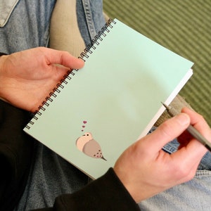 Puede incluir: Una libreta de espiral azul claro con una ilustración de un pájaro blanco y gris y corazones rosas. La libreta está abierta y una persona sostiene un bolígrafo plateado.