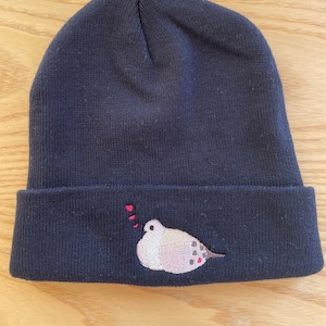 May include: A dark blue knitted beanie with a small embroidered bird design. The bird is beige with brown spots and has a red heart above its head.