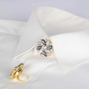 May include: A gold-toned collar clip with clear and dark blue crystals. The clip is on a white shirt collar.