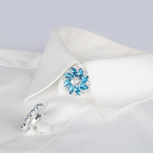 May include: A blue and silver rhinestone collar clip in the shape of a flower. The clip is on a white shirt collar.