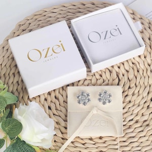Peut inclure: Deux boîtes blanches avec le logo "Ozei Jewelry" imprimé en or. Les boîtes sont sur une surface en paille tressée. Une petite pochette blanche avec le logo "Ozei" imprimé en or est sur la surface en paille. La pochette contient deux boucles d'oreilles en argent en forme de fleur avec des cristaux transparents.