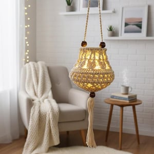 Peut inclure: Une lanterne suspendue en crochet de couleur crème avec une lumière chaude et brillante à l'intérieur. La lanterne est suspendue par une corde crème avec des perles en bois. Une gland crème pend du bas. La lanterne est dans une pièce chaleureuse et bien éclairée.