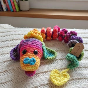 Può includere: Un giocattolo per cani all'uncinetto colorato con testa, orecchie e corpo color arcobaleno. Il giocattolo ha un papillon blu, occhi neri e un corpo a spirale nei toni dell'arancione, del rosa e del viola. Un pezzo a forma di cuore giallo pende dall'estremità.