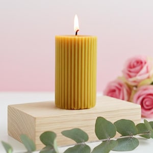 May include: A lit, yellow, ribbed pillar candle sits on a small, natural wood block. The candle is in focus, with a soft pink background and a few pink roses. Green eucalyptus leaves are in the foreground, adding a natural touch to the scene.