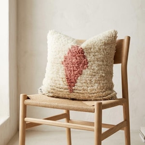 May include: A cream-colored, textured pillow with a pink diamond design rests on a light wood chair with a woven seat. The pillow is the focal point, showcasing a soft, inviting texture. The chair is in a neutral-toned room.