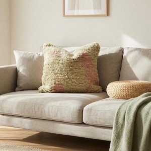 May include: A light gray sofa with a textured, olive green and pink pillow. A woven, round ottoman and a sage green throw blanket are also on the sofa. The room has a neutral color palette.