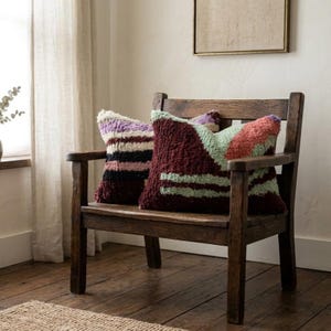 Peut inclure: Deux coussins décoratifs reposent sur un banc en bois. L'un présente un motif à rayures noires, violettes et crème. L'autre a un design géométrique bordeaux, vert et pêche. Le banc est en bois foncé.