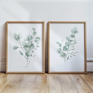 May include: Two rectangular wall art prints in light wood frames, each featuring a watercolour illustration of green eucalyptus branches with rounded leaves on a white background, displayed side by side on a wooden floor surface.