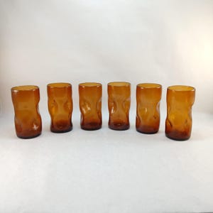 May include: Six amber-colored drinking glasses. Each glass has a unique, textured, organic shape. The glasses are cylindrical with a slightly irregular surface. The color is a warm, transparent amber, suggesting they are made of glass.