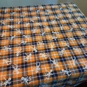May include: Orange and black plaid fabric with a repeating pattern of white skeletons. The fabric is arranged on a table, showcasing the Halloween-themed design. The plaid pattern is composed of orange, black, and white lines.