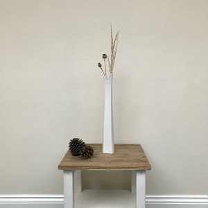 May include: White ceramic vase with a unique, asymmetrical opening, holding dried grasses and seed pods. The vase is on a white wooden table with a brown top, and there are two pine cones on the table.