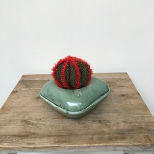 May include: A green ceramic pillow with a red and green knitted cactus on top. The pillow and cactus are sitting on a wooden table.