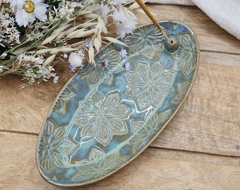 Könnte beinhalten: Ein tealblauer Keramik-Räucherstäbchenhalter mit floralem Design. Der Halter ist oval geformt und hat ein kleines Loch in der Mitte für Räucherstäbchen.