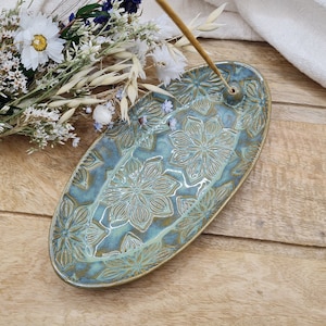 Könnte beinhalten: Ein tealblauer Keramik-Räucherstäbchenhalter mit floralem Design. Der Halter ist oval geformt und hat ein kleines Loch in der Mitte für Räucherstäbchen.