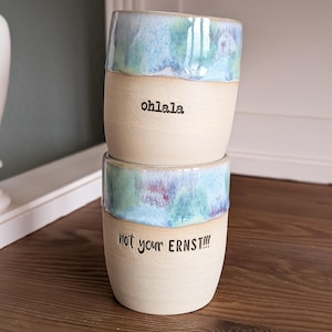 Könnte beinhalten: Zwei gestapelte Keramikbecher mit matter beiger Basis und einem glänzend gesprenkelten blau-grünen Glasurrand. Der obere Becher trägt den schwarzen Schriftzug "ohlala." und der untere Becher den schwarzen Schriftzug "not your ERNST!!!" in Schreibmaschinenschrift.