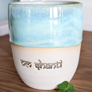 K&ouml;nnte beinhalten: Eine Keramiktasse mit zweifarbigem Design. Die obere H&auml;lfte ist hellblau mit Marmoreffekt, die untere H&auml;lfte ist cremefarben. Die Worte "Om Shanti" sind in den cremefarbenen Bereich ge&auml;tzt. Ein gr&uuml;nes Kleeblatt liegt neben der Tasse.