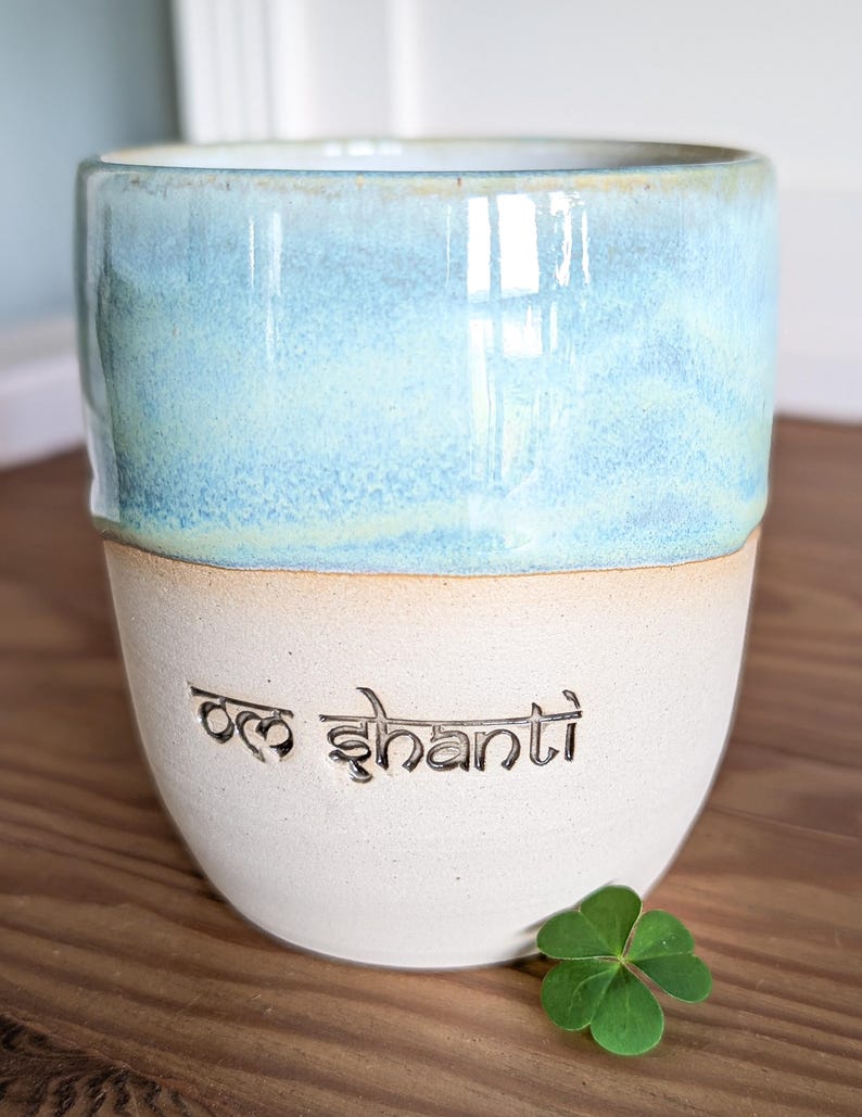 K&ouml;nnte beinhalten: Eine Keramiktasse mit zweifarbigem Design. Die obere H&auml;lfte ist hellblau mit Marmoreffekt, die untere H&auml;lfte ist cremefarben. Die Worte "Om Shanti" sind in den cremefarbenen Bereich ge&auml;tzt. Ein gr&uuml;nes Kleeblatt liegt neben der Tasse.