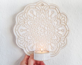 May include: White ceramic wall decor with a raised mandala design and a small candle holder. The candle holder is attached to the bottom of the mandala and has a white ceramic candle inside.