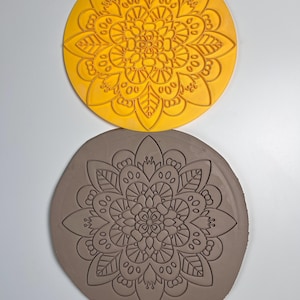 Könnte beinhalten: Ein gelber, kreisförmiger Tonstempel mit einem detaillierten floralen Mandala-Design. Darunter ein brauner Tonabdruck desselben Mandalas. Der Stempel wird verwendet, um dekorative Muster in Ton oder anderen weichen Materialien zu erstellen. Das Design zeigt Blütenblätter und Blätter.