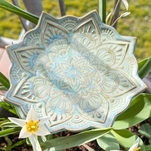 Könnte beinhalten: Eine dekorative, sternförmige Keramikschale mit detailliertem Mandala-Muster. Die Schale hat eine Mischung aus Blau-, Türkis- und Goldtönen. Die Ränder sind leicht erhöht, und das Design ist geprägt. Die Schale ist von grünen Blättern und weißen Blumen umgeben.
