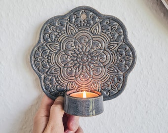 May include: A gray ceramic wall sconce with a mandala design and a candle holder. The candle is lit and glowing.