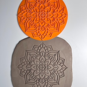 Könnte beinhalten: Ein orangefarbener, kreisförmiger Mandala-Stempel liegt über einem Tonabdruck desselben Designs. Der Stempel weist ein detailliertes, geschichtetes Blumenmuster auf. Der Tonabdruck ist hellbraun und zeigt das gestempelte Mandala-Design.